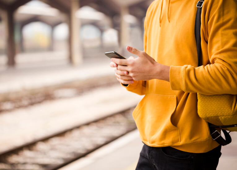 Zdjęcie przedstawiające osobę stojącą na dworcu kolejowym która korzysta z telefonu komórkowego